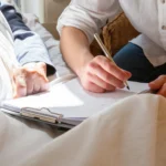 A caregiver sitting beside an elderly patient’s bed, holding a pen and helping the person fill out documents on a clipboard.