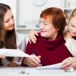 ثلاثة أجيال من النساء يبتسمن فوق أوراق على طاولة، إحداهن تكتب بقلم أزرق.